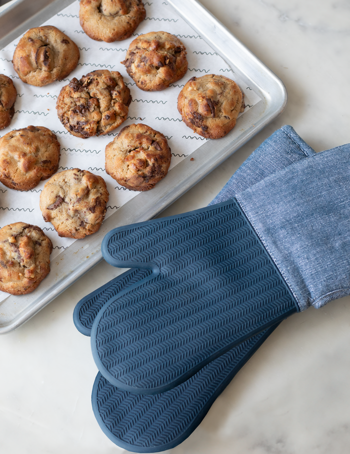 A pair of blue silicone oven mitt with a textured grip and a light blue fabric cuff, displayed next to a baking sheet with cookies.