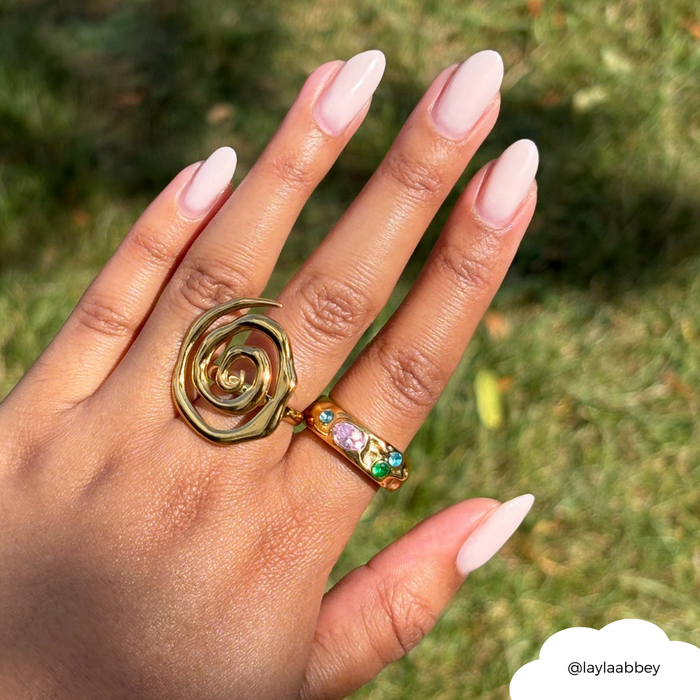 Hand with long nude nails wears a gold Sunset Spiral Ring and another gold ring, shown against green grass.