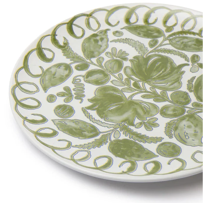 A close-up of a white ceramic plate decorated with a hand-painted green botanical pattern of leaves, flowers, and scrolling borders, set against a plain white background.