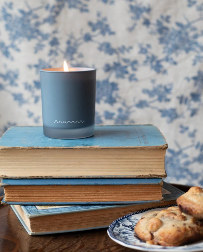 Blue Apotheke candle with white “Maman” logo on a stack of books, with cookies on a plate, against a blue-and-white patterned wall.