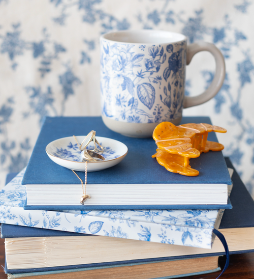 Blue and white floral mug next to the croissant hair clip and the blue and white floral jewelry dish, featuring the croissant necklace and a ring inside. Items placed on top of a stack of blue and white floral notebooks.