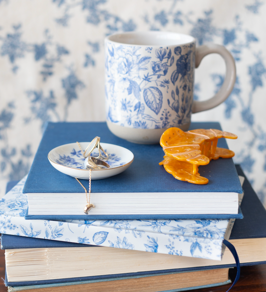 Blue and white floral mug next to the croissant hair clip and the blue and white floral jewelry dish, featuring the croissant necklace and a ring inside. Items placed on top of a stack of blue and white floral notebooks.