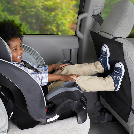 A young child smiles in a car seat, their feet resting on the Car Seat Kick Mat (2 Pack), which protects the front seat back from dirt and scuffs.