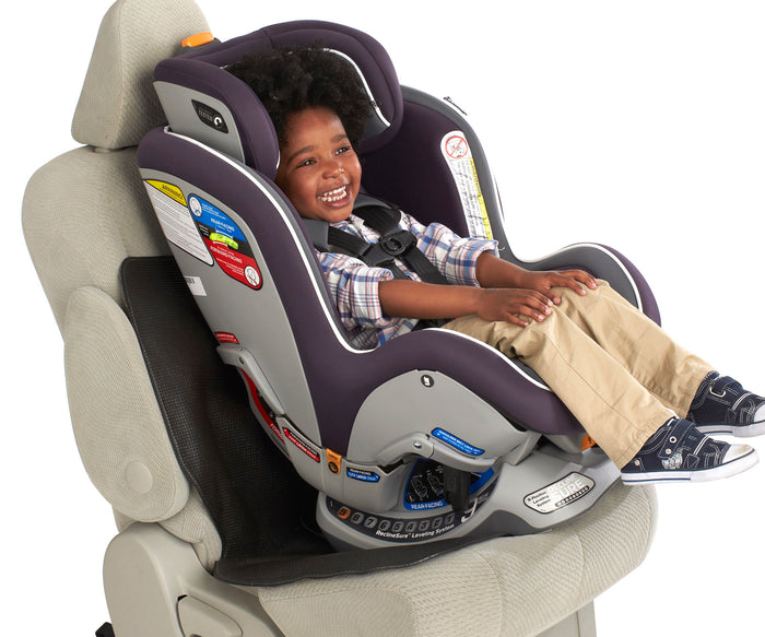 A young child smiles while sitting in a rear-facing car seat, securely fastened with the Car Seat Undermat underneath. The child wears a plaid shirt and khaki pants. The car seat has safety labels and comfortable padding.