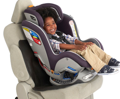 A young child smiles while sitting in a rear-facing car seat, securely fastened with the Car Seat Undermat underneath. The child wears a plaid shirt and khaki pants. The car seat has safety labels and comfortable padding.