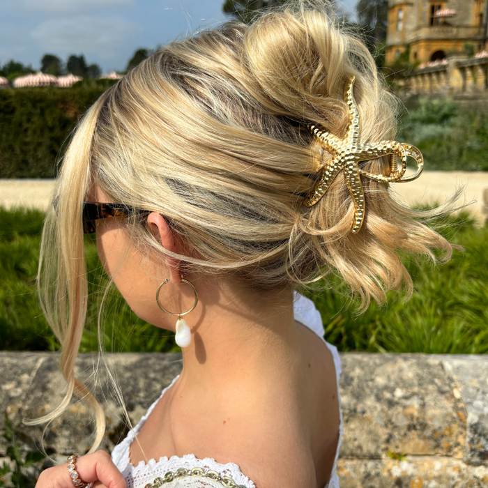 Woman with a messy blonde bun wears a gold metal starfish claw clip from Coconut Lane, sunglasses, and a pearl earring.