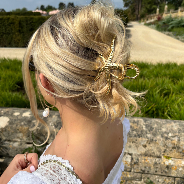 Blonde woman in a loose updo held by a gold metal starfish claw clip, standing outside in a garden.