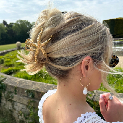 Blonde hair in an updo with a gold metal starfish claw clip, paired with gold hoops and pearl drop earrings.