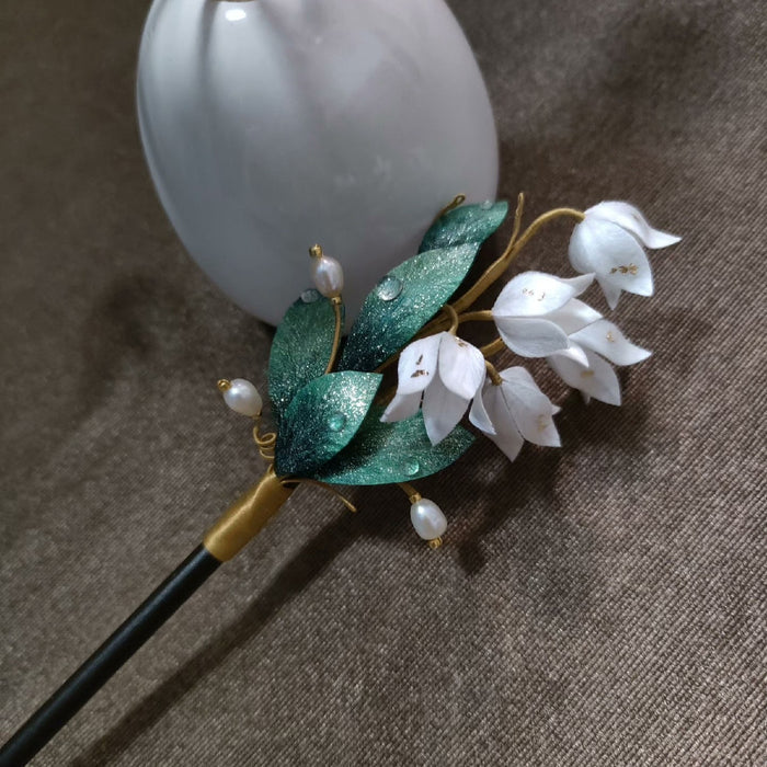 Ronghua handmade lily of the valley hairpin with white silk bell flowers, deep green glitter leaves with dew drops, pearl accents, and a dark wooden pin, displayed against a white ceramic vase on a grey fabric background