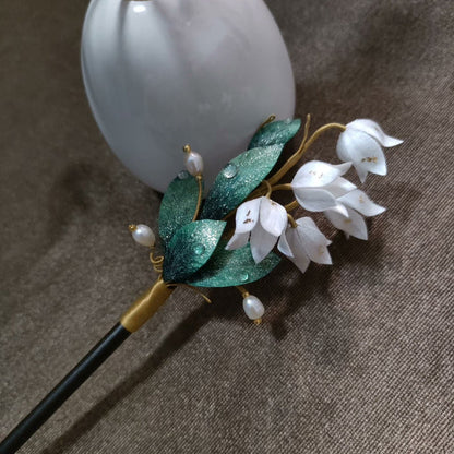 Ronghua handmade lily of the valley hairpin with white silk bell flowers, deep green glitter leaves with dew drops, pearl accents, and a dark wooden pin, displayed against a white ceramic vase on a grey fabric background