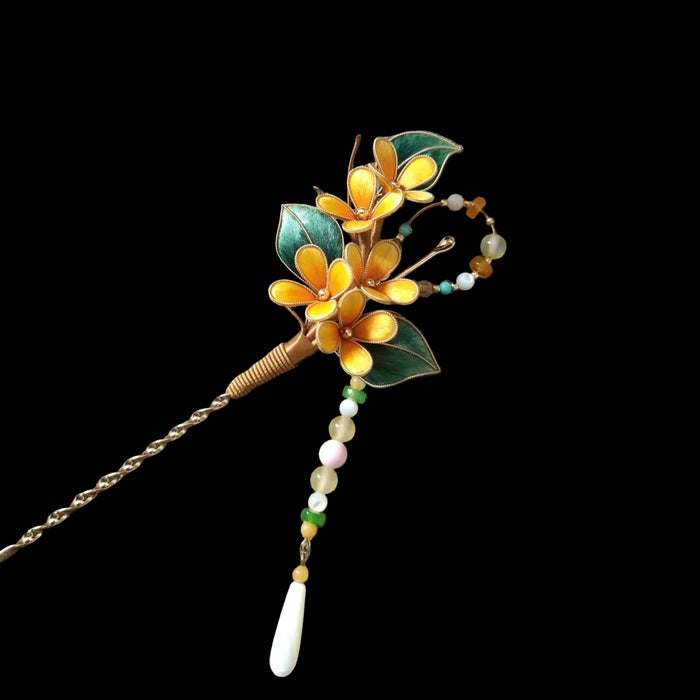 Ronghua handmade osmanthus hairpin with three golden yellow silk flowers, gold edge trim, dark green leaves, copper-wrapped stem, and long beaded chain drop ending in a white shell pendant, displayed on dark grey fabric