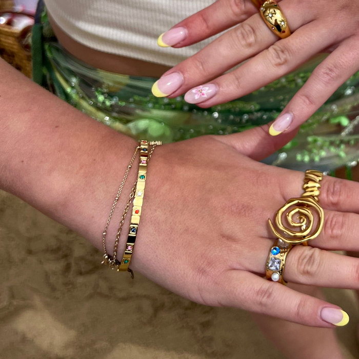 Hands with colorful nail art, gold rings, bracelets, and a gold Coconut Lane Jewelbeam Bangle against a green sleeve.