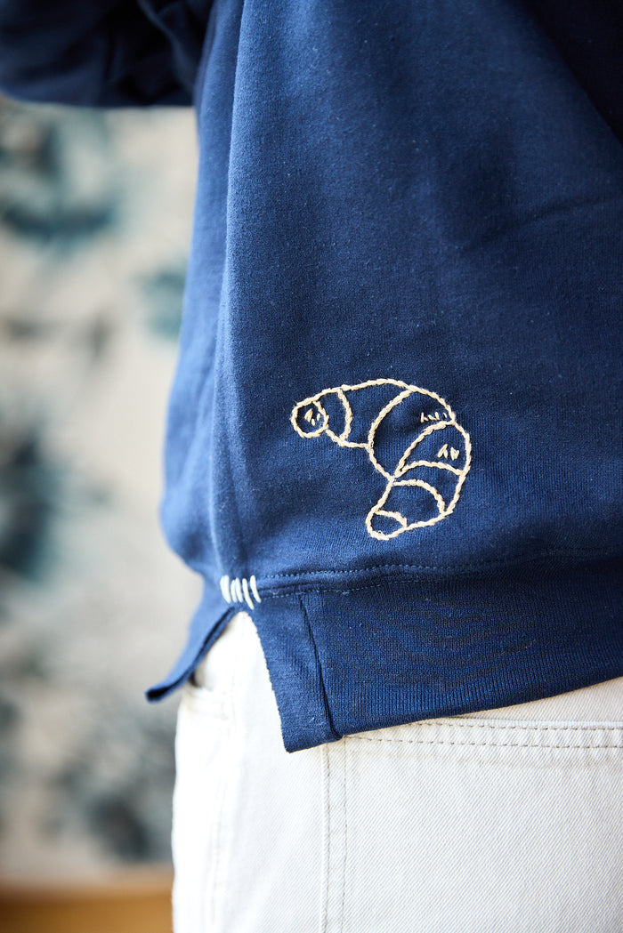 A navy blue sweatshirt with a beige hand-embroidered croissant at the hem.