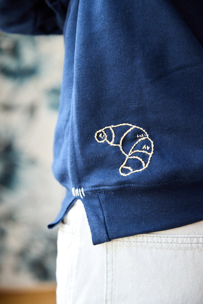 A navy blue sweatshirt with a beige hand-embroidered croissant at the hem.