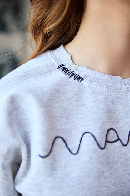 A grey sweatshirt with blue hand-embroidered "maman" logo on the front and lines details on the collar.