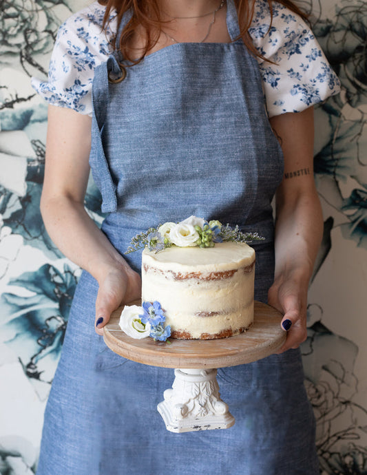 a person wearing a blue chambray baker's apron holding a cake.