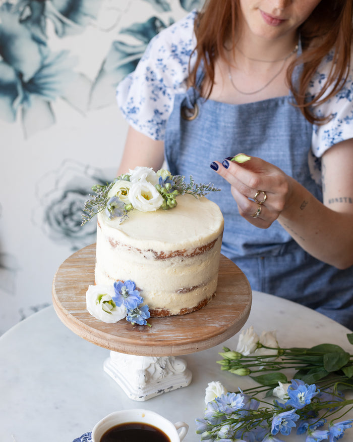 a person wearing a blue chambray baker's apron decorating a cake.