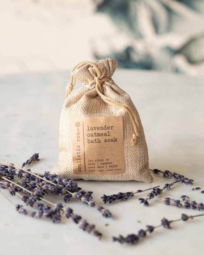 Small burlap bag labeled 'lavender oatmeal bath soak' is displayed with scattered lavender sprigs on a light background.