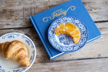 Gold croissant-shaped hair clip inspired by French pastry, on a small plate next to a real croissant.