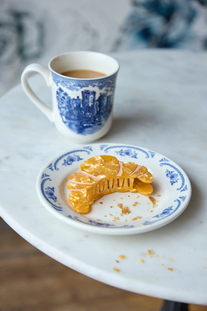 Gold croissant-shaped hair clip inspired by French pastry, on a small plate.