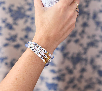Three bracelets worn on wrist, each with a gold croissant bead and letter beads spelling a different word “bonjour”, “maman”, and “je t’aime” in shades of blue, blue-and-white, and white-and-gold beads.