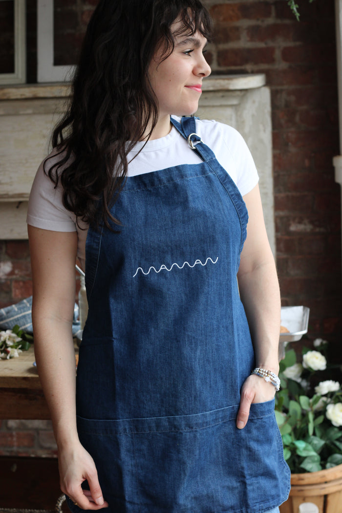Person wearing a blue denim apron with pockets and the “Maman” logo embroidered on it.