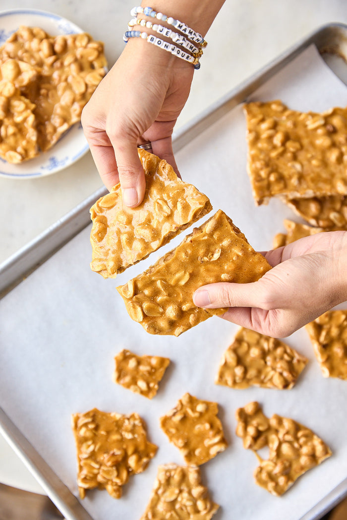Hands breaking a piece of peanut brittle, with more pieces on a baking sheet.