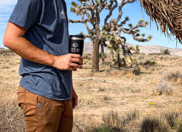 Man in outdoor clothing holding a black travel mug in a desert landscape with Joshua trees.