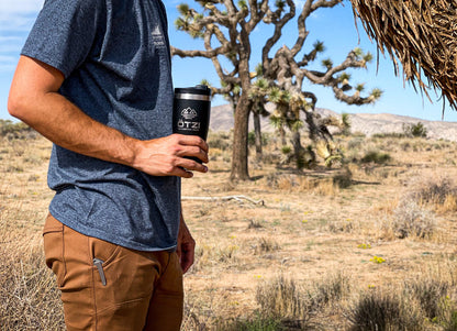 Man in outdoor clothing holding a black travel mug in a desert landscape with Joshua trees.