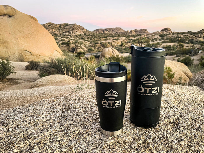 Two Ötzi travel mugs on a rock in a desert landscape at sunset