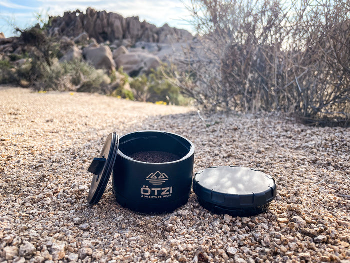 Black OTZI container with open lid showing ground coffee on rocky desert terrain.