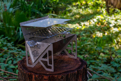 Stainless steel portable grill set up on a tree stump in a forested area.