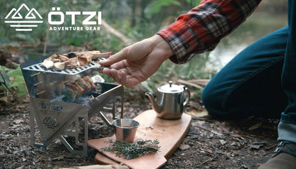 Person grilling food on a compact stainless steel camping stove beside a teapot and herbs outdoors.