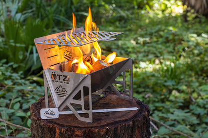 Compact stainless steel campfire stove with open flame set on a tree stump in a forest.