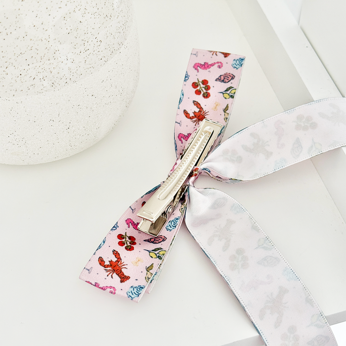 Coconut Lane Cococabana ribbon bow clip on a white surface beside a speckled round object.