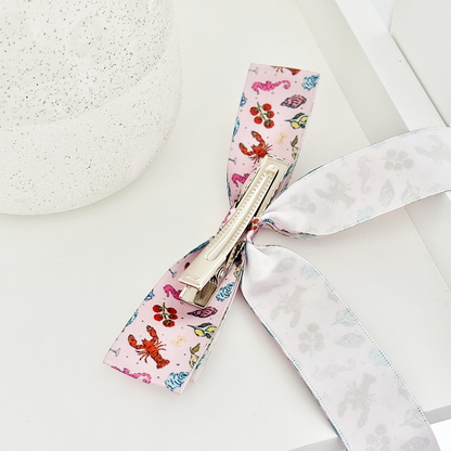 Coconut Lane Cococabana ribbon bow clip on a white surface beside a speckled round object.