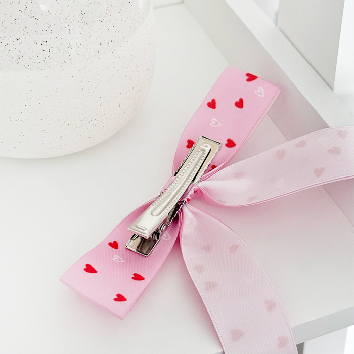 Pink ribbon bow hair clip with a hearts pattern, placed on a white surface beside a speckled white container.
