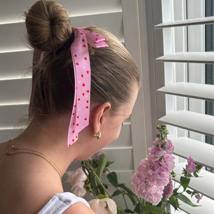 Woman wears Coconut Lane Ribbon Bow Clip with a cute heart pattern, standing by pink flowers and white window blinds.