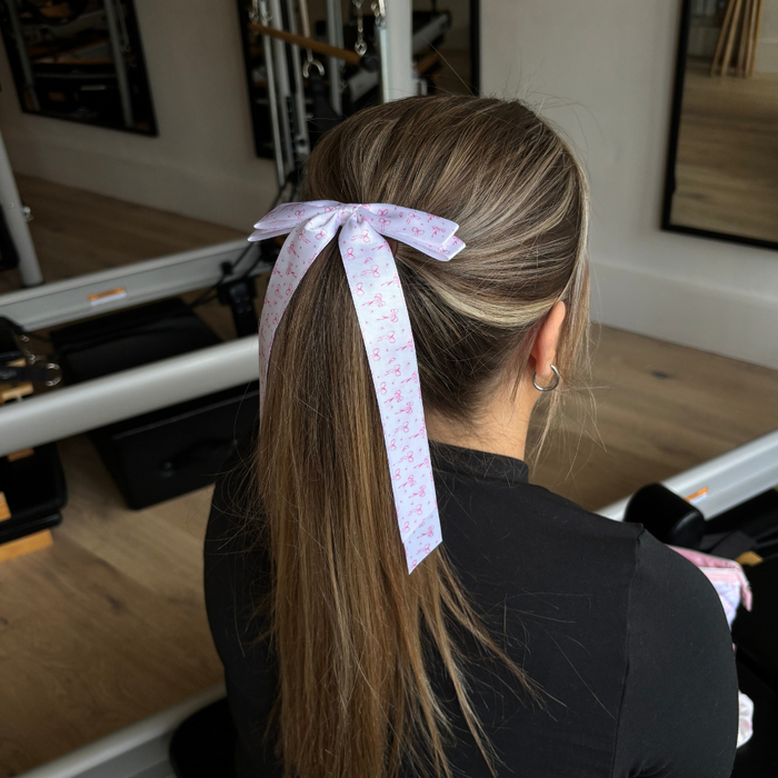 Woman with long hair wearing a pale pink Coconut Lane ribbon bow clip, seated in a gym.