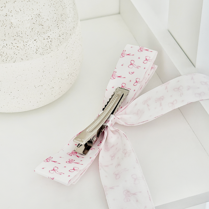 White ribbon bow hair clip with pink designs, shown next to a speckled container.