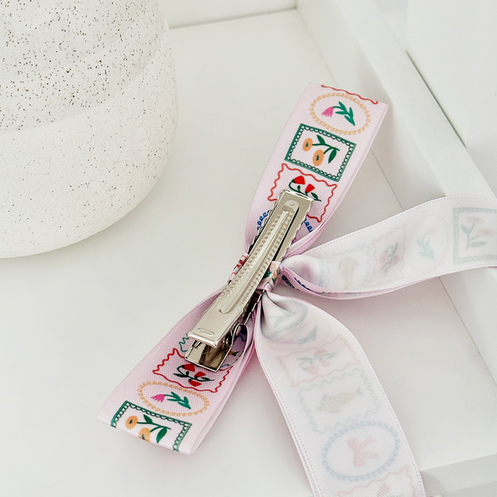 Cream ribbon bow hair clip next to a white speckled container on a white surface.