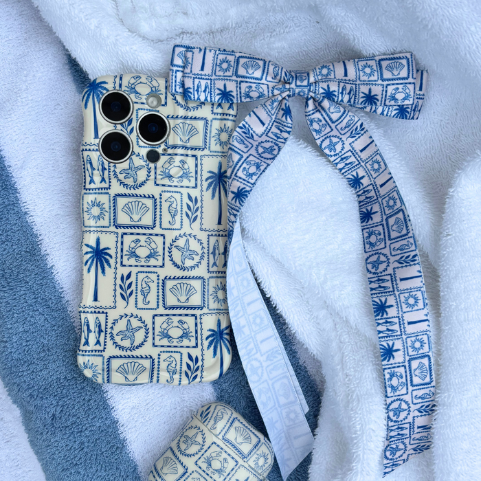 Blue and cream seashell-pattern phone case, bow, and earbud case set with ribbon bow clip on a white towel.