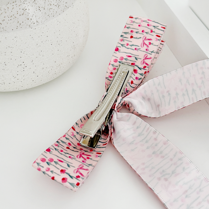 Peony pink ribbon bow hair clip from Coconut Lane on a white surface next to a speckled container.