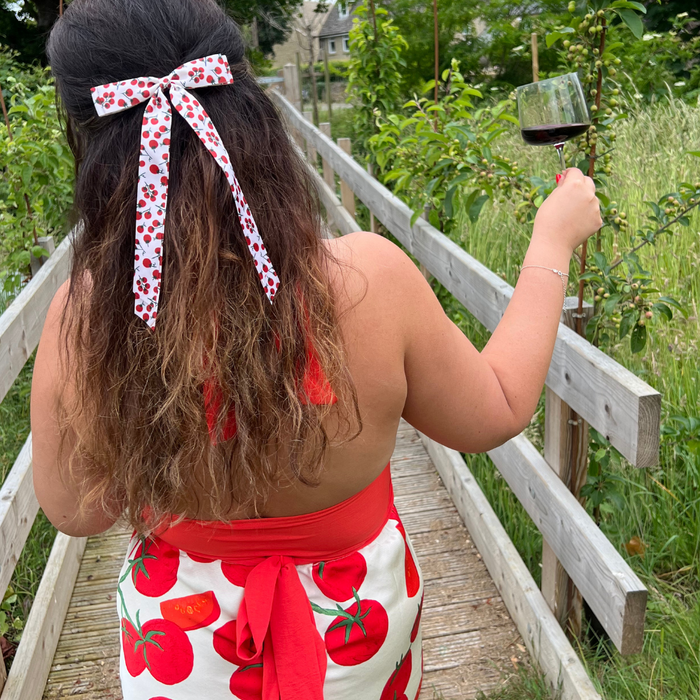 Woman in a tomato-print dress wears a red ribbon bow hair clip, holding red wine on a wooden path.
