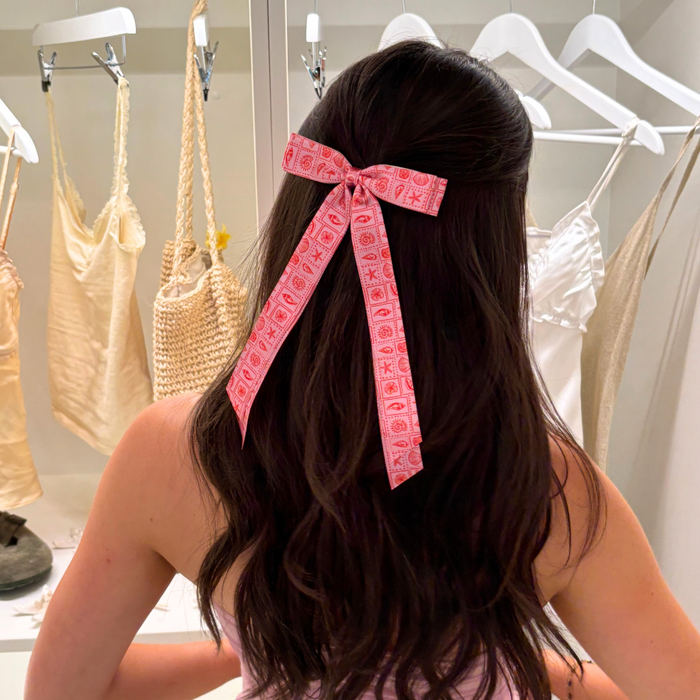 Woman with long brown hair wears a white ribbon bow clip while standing by hanging dresses and woven bags.