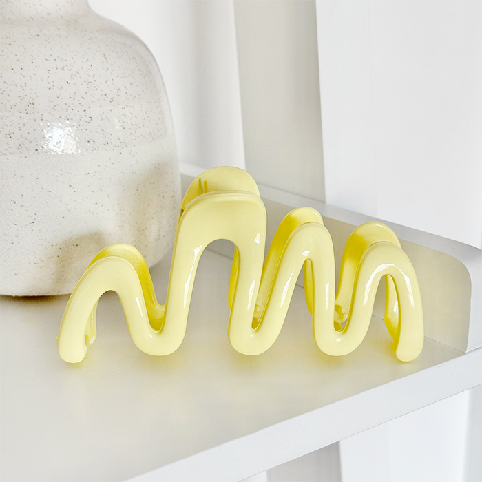 Glossy yellow Coconut Lane Squiggle Claw Clip on a white shelf next to a speckled white vase.
