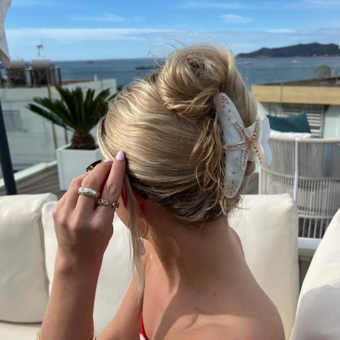 Blonde woman with hair in a bun, secured by a golden starfish claw clip, sits near the ocean.