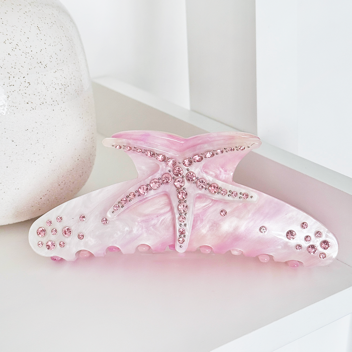 Pink starfish-shaped Coconut Lane claw clip with rhinestones on a white surface beside a ceramic vase.