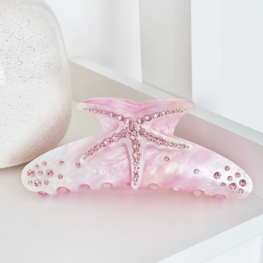Pink starfish-shaped Coconut Lane claw clip with rhinestones on a white surface beside a ceramic vase.