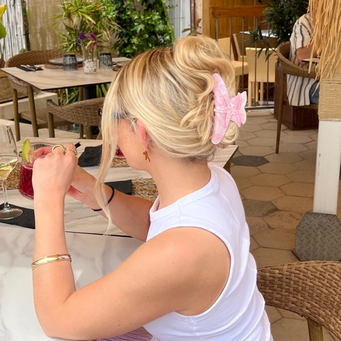 Blonde woman with hair in a bun wears a pink starfish claw clip while sitting at an outdoor café holding a drink.